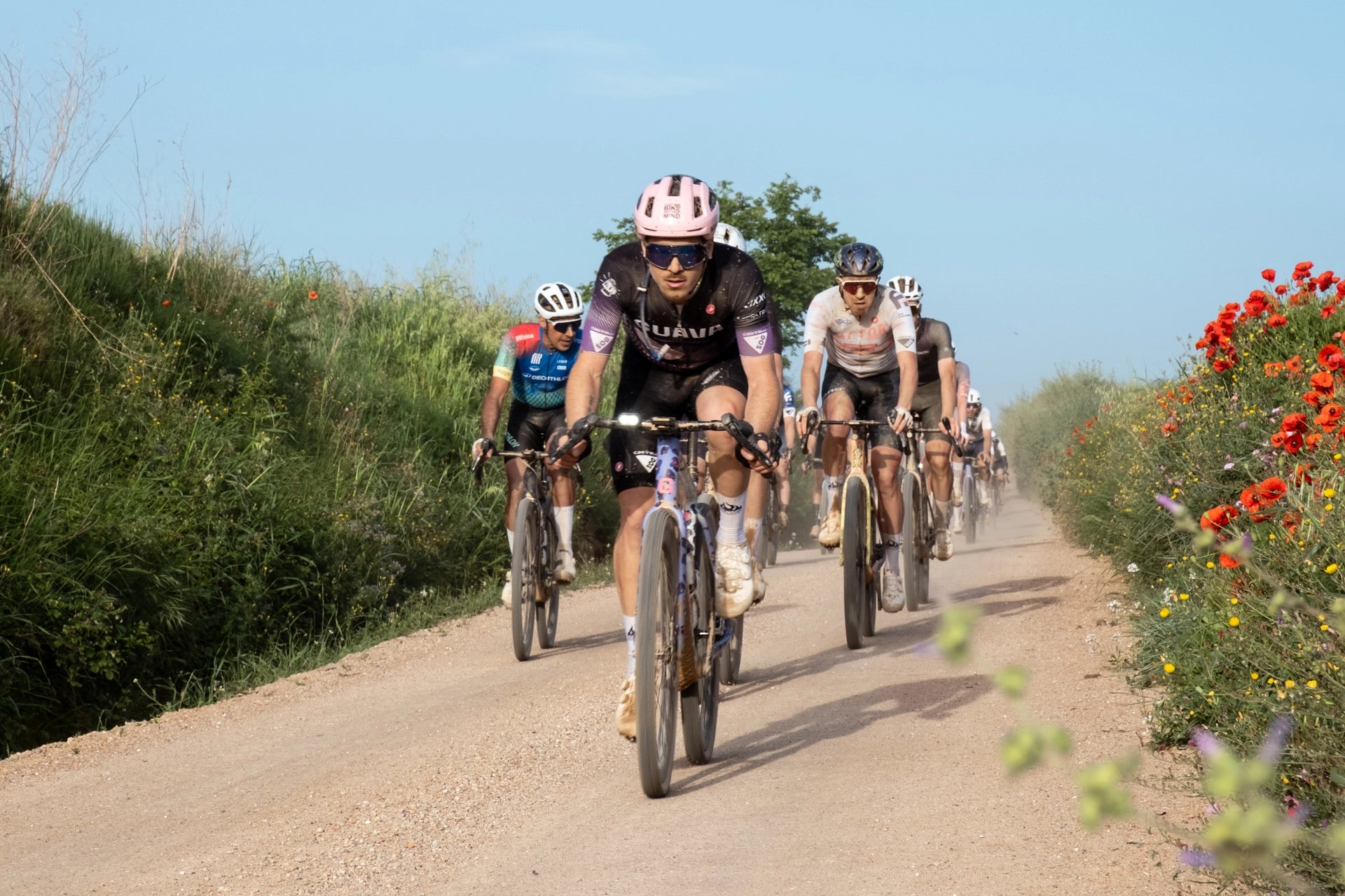 grupo de ciclistas en el campo, ciclista con casco rosa, mallot lila y negro.