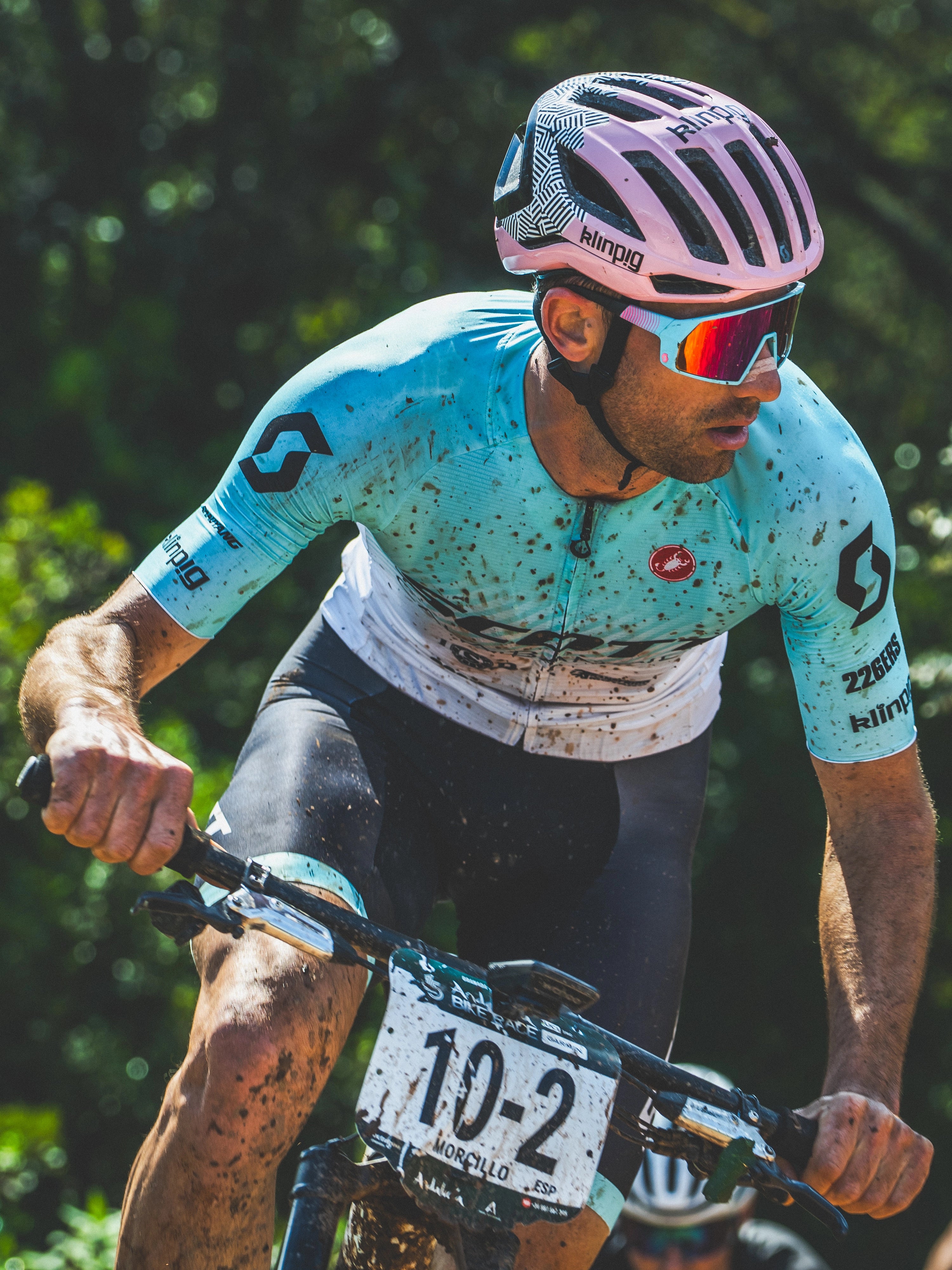 casco rosa lentes de ciclismo corrdor con la pierna negra de barro maillot azul y blanco 