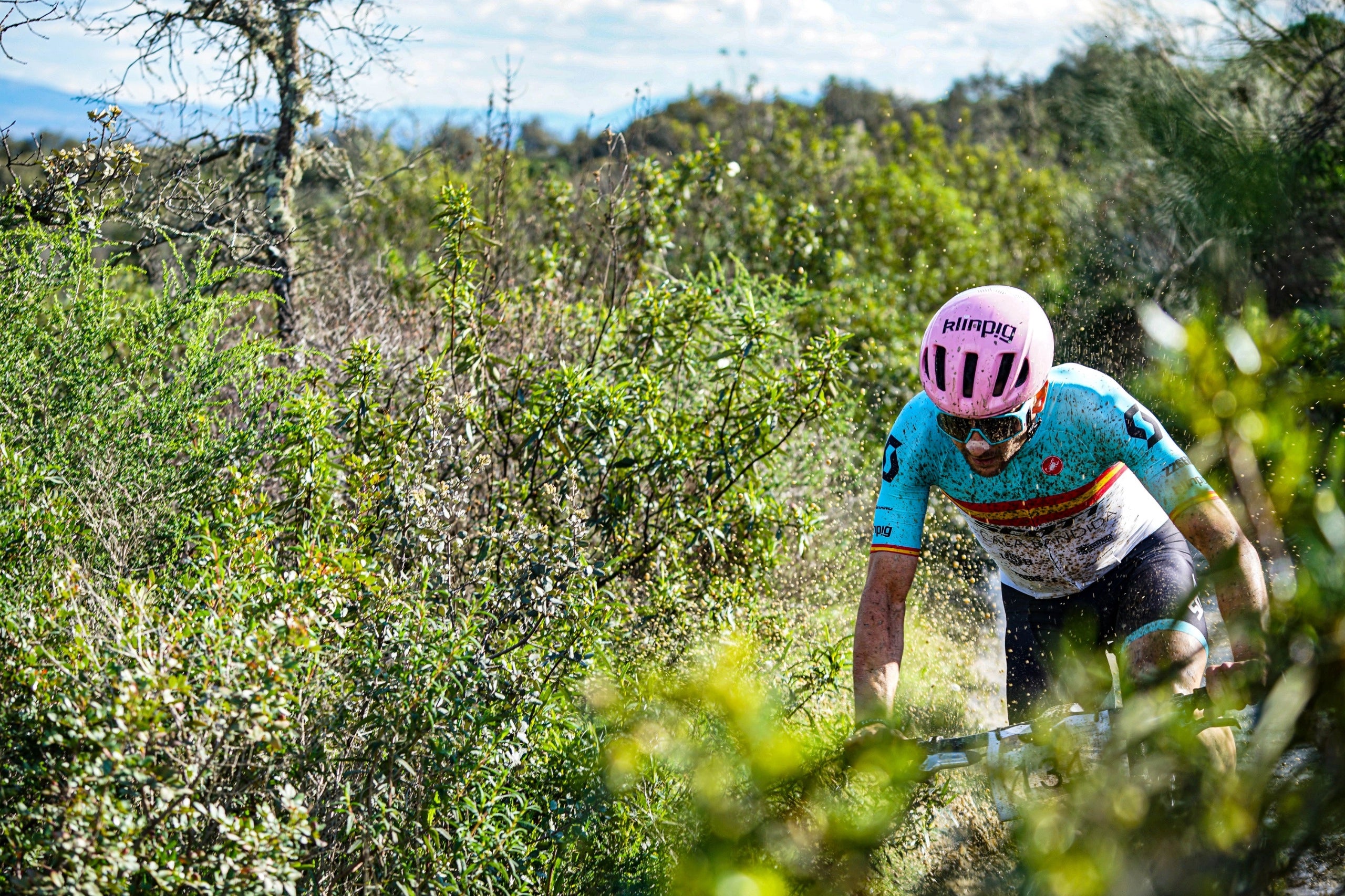 Ciclista descendiendo con técnica en un camino estrecho de montaña rodeado de vegetación densa y árboles bajos, montando una bicicleta gravel y vistiendo maillot claro con casco rosa ventilado